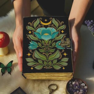 Puede incluir: Un diario negro con un diseño floral turquesa, hojas verdes y detalles dorados. El diario tiene un cierre de broche y bordes dorados. La portada también presenta una luna creciente y estrellas.