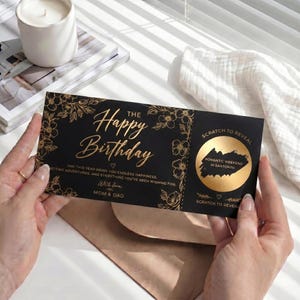 May include: A black birthday scratch-off card with gold floral accents and text that reads "Happy Birthday." The card includes the message "Romantic Weekend in Santorini" and is held by a person.