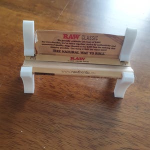 May include: A small white bench designed to hold RAW Classic rolling papers. The bench features a minimalist design with four legs and two back supports. The papers display the text "RAW CLASSIC" and "THE NATURAL WAY TO ROLL".