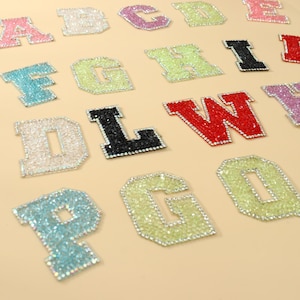 Puede incluir: Una colección de parches con letras del alfabeto estilo universitario cubiertas de brillantes pedrería. Las letras aparecen en varios colores, incluyendo azul claro, verde lima, negro, rojo, rosa y transparente, cada una con un borde plateado.