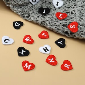 Op de afbeelding: Een verzameling kleine hartvormige geborduurde patches in rood, zwart en wit, elk voorzien van een enkele hoofdletter. De patches liggen verspreid over een oppervlak, waarbij sommige zijn bevestigd aan een grijze gehaakte stof. De letters omvatten C, D, W, X, T, H, M, N, R, Y, S, E, J en U.