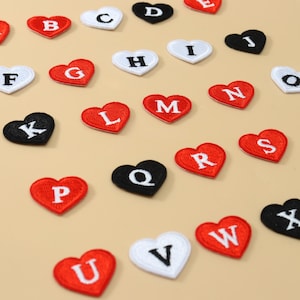 May include: A collection of heart-shaped embroidered patches in red, white, and black. Each patch features a single capital letter of the alphabet stitched in a contrasting color. The patches are arranged in a scattered pattern across the frame.