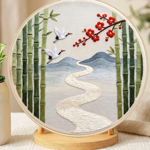 Op de afbeelding: Een ronde borduurring met een stoffen paneel met een landschap. Het ontwerp toont groene bamboestengels, twee witte kraanvogels in de vlucht, een kronkelende witte rivier, blauwe bergen en een tak met helderrode bloemen.