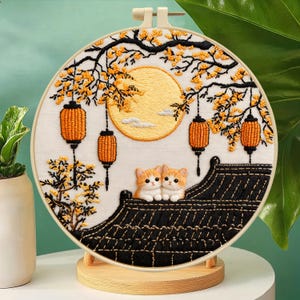May include: A circular embroidery hoop featuring a stitched scene of two orange kittens sitting on a black tiled roof. Above them, a large yellow moon hangs among tree branches with yellow blossoms and three hanging orange lanterns on black cords.