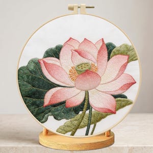 May include: Embroidered lotus flower art in a round wooden hoop. The flower features pink petals with white accents, a yellow center, and green leaves. The artwork is displayed on a wooden stand, showcasing the detailed needlework.