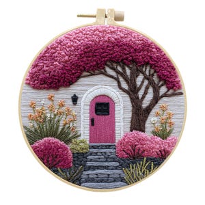 Op de afbeelding: Een ronde borduurring met een borduurwerk van een roze deur met een stenen pad, omringd door gestructureerde roze en magentakleurige struiken, een grote boom met dicht roze gebladerte en kleine oranje bloemen op een lichtgrijze achtergrond.