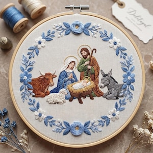May include: A circular wooden embroidery hoop containing a fabric panel with a hand-stitched nativity scene. The design features a central manger with a baby, surrounded by figures, a brown ox, a white lamb, and a gray donkey, framed by blue flowers.