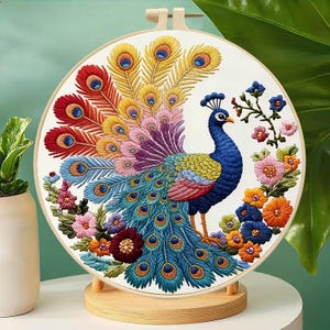 May include: A circular embroidery hoop containing a colorful peacock design stitched on white fabric. The peacock features a vibrant, fanned tail with red, yellow, purple, and blue feathers, surrounded by embroidered flowers in various shades.