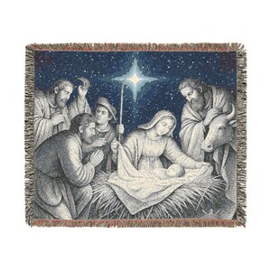 May include: A woven throw blanket featuring a nativity scene with a central star, figures gathered around a manger, and a cow, rendered in shades of blue, cream, and brown with a fringed edge. The design depicts a starry night sky background.