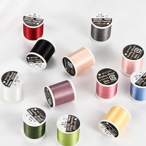 Puede incluir: Una colección de coloridas bobinas de hilo para abalorios Miyuki en varios tonos, incluyendo rojo, negro y rosa. Cada bobina está etiquetada con detalles como el tipo de hilo, el número de lote y el código de color. Los hilos son ideales para trabajos con abalorios y la creación de joyas.