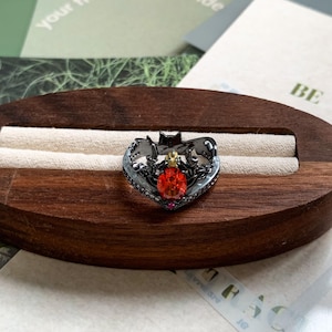 May include: A black metal ring featuring a central oval-shaped orange gemstone, a small clear stone above it, and a tiny pink stone at the base. The band is shaped like a bat with wings extending outward, accented with small clear stones along the edges.