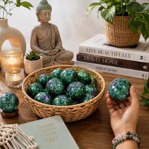 Può includere: Una collezione di pietre sferiche con finitura lucida, caratterizzate da un mix vibrante di motivi maculati verde intenso, smeraldo e viola. Queste pietre rotonde sono esposte in un cesto di vimini intrecciato, con una tenuta in mano.