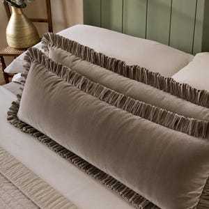 Puede incluir: Un cojín alargado de forma rectangular hecho de tela de terciopelo color topo. El cojín presenta un borde decorativo con volantes fruncidos en los extremos superior e inferior, creando un borde texturizado que enmarca la superficie suave.