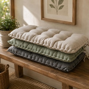May include: A stack of three rectangular tufted bench cushions in beige, sage green, and charcoal gray. Each cushion features a textured fabric cover with visible stitched seams along the edges and a series of tufted indentations on the top surface.