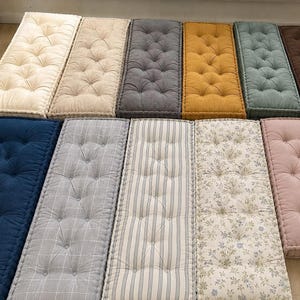 Op de afbeelding: Een verzameling rechthoekige vloerkussens in verschillende kleuren en patronen. De kussens zijn voorzien van knopen en omvatten tinten crème, grijs, mosterdgeel, blauw en roze. Sommige hebben streep- of bloemmotieven, andere zijn effen.