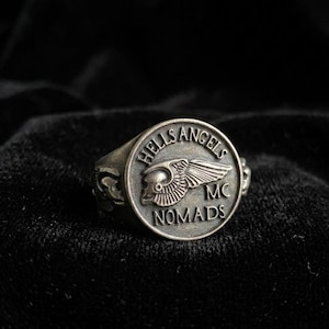 Op de afbeelding: Een zilverkleurige metalen ring met een cirkelvormig voorvlak met een reliëf van een gevleugelde schedel. De tekst HELLS ANGELS MC NOMADS is gegraveerd rond de omtrek van het cirkelvormige vlak. De ring heeft decoratieve details aan de zijkanten.