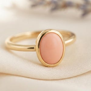 Può includere: Un anello in oro con una pietra preziosa cabochon di forma ovale color pesca chiaro, incastonata in una lunetta in oro lucido. La fascia è un semplice filo d'oro arrotondato che si collega all'incastonatura centrale, creando un gioiello.