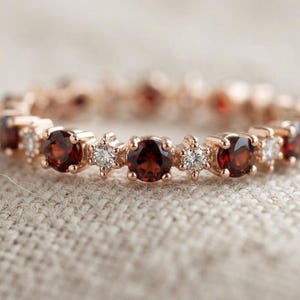 Puede incluir: Una alianza de oro rosa con un patrón alterno de piedras preciosas de granate rojo oscuro redondas y diamantes blancos transparentes más pequeños, dispuestos en un diseño circular continuo que rodea toda la banda del anillo de metal.