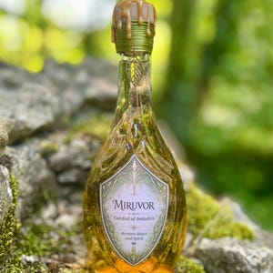 Peut inclure: Une bouteille en verre transparent remplie d'un liquide doré, avec un bouchon en liège scellé par de la cire beige coulante et entouré de fil vert. Une breloque en métal en forme de feuille pend au goulot. L'étiquette indique Miruvor Cordial of Imladris.