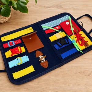 May include: A navy blue sensory activity board featuring various fasteners including a yellow zip, red and yellow buckles, a brown snap-button pocket, a toggle closure, blue and yellow straps, a red lace-up section, and a light blue polka dot strip.
