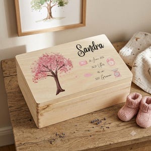 Könnte beinhalten: Eine rechteckige Aufbewahrungsbox aus hellem Holz mit einem aufgedruckten rosa Kirschblütenbaum und dem Namen Sandra. Der Deckel zeigt Geburtsdaten: 26 June 2026, 06:25 Uhr, 46 cm und 3255 Gramm, ergänzt durch kleine rosa Symbole für Kalender, Uhr und Waage.