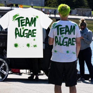 May include: A white short-sleeve t-shirt featuring the text "TEAM ALGAE" printed in a dark green, dripping, paint-like font. The design includes scattered green paint splatter accents across the front and back of the garment fabric.
