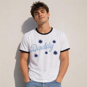 Puede incluir: Camiseta blanca con cuello redondo y ribete negro. La palabra "Daddy" en azul claro, rodeada de estrellas azules oscuras. Estilo casual.