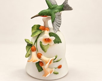 May include: A white porcelain bell featuring a sculpted green and grey hummingbird perched on top. The bell is decorated with three-dimensional orange and white trumpet-shaped flowers and green leaves attached to the side of the smooth white body.