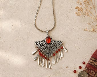 Può includere: Una collana color argento con un ciondolo triangolare caratterizzato da intricati motivi a volute. Il ciondolo è centrato da una pietra rossa ovale e decorato con molteplici ciondoli argentati pendenti e piccole perline rosse sul bordo.