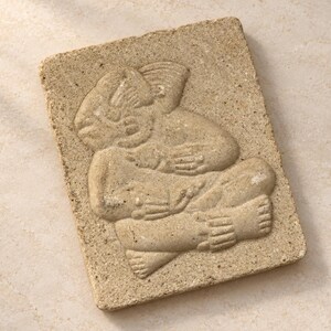 May include: A rectangular beige stone plaque featuring a relief carving of a seated figure with stylized features. The figure is depicted in a cross-legged position with hands resting on the chest and lap, showcasing intricate textures and details.