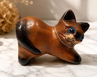 May include: A small, hand-carved wooden cat figurine with a smooth, polished brown finish. The cat features dark brown accents on its ears, face, and tail, bright blue painted eyes, and a thin silver-toned metal chain necklace around its neck.