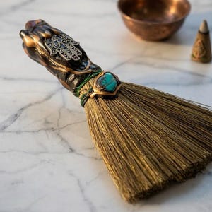 May include: A decorative broom with a bronze-colored handle featuring a Hamsa hand design and a turquoise stone accent. The broom's bristles are a golden-brown color. A copper bowl and incense cone are in the background.