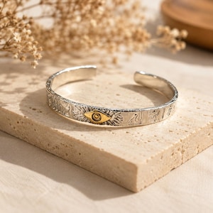Könnte beinhalten: Ein silberner Armreif mit texturierter Oberfläche, gravierten Symbolen und einem zentralen goldfarbenen Augenmotiv. Das offene Armband hat eine polierte Oberfläche und zeigt verschiedene eingravierte Muster entlang des äußeren Umfangs.
