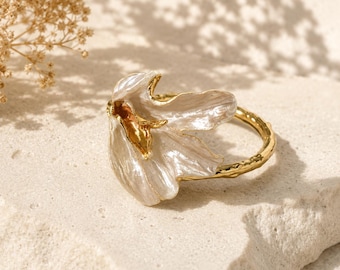 May include: A gold-toned ring featuring a large, iridescent white flower-shaped centerpiece with delicate, wavy petals. The band of the ring has a textured, branch-like appearance, providing a natural aesthetic to the overall jewelry piece.