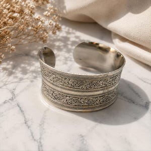 May include: A wide silver-toned cuff bracelet featuring an open back design. The exterior surface is decorated with two horizontal bands of intricate, raised filigree scrollwork patterns separated by a smooth, plain central silver-toned metal strip.