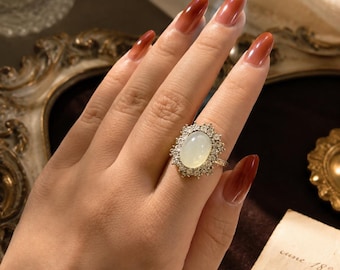 May include: A gold ring featuring a large, oval-shaped, milky white cabochon stone at the center, surrounded by a halo of small, clear, sparkling stones set in a decorative, textured metal frame that extends outward in a sunburst pattern.