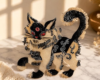 May include: A flat, decorative cat figurine featuring a black and cream color scheme with intricate swirling patterns. The cat has three red eyes, including one on its forehead, and a long, curved tail. The design is detailed with fine lines.