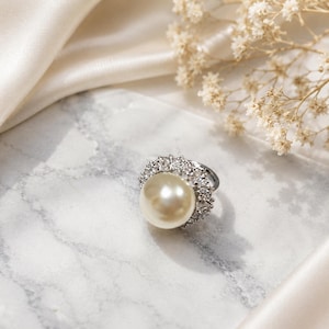 Puede incluir: Un anillo de tono plateado con una perla grande, redonda y de color crema en el centro, rodeada por un halo de pequeñas piedras claras y brillantes. El anillo tiene una banda de metal pulido y un engaste detallado que enmarca la perla.