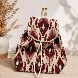 May include: A backpack featuring a cream-colored fabric base with a repeating geometric pattern in deep red and dark gray. The bag includes a top flap with a single metal snap closure, a top handle, and two off-white drawstring cords for closure.