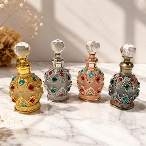 May include: Four small decorative perfume bottles with ornate metal filigree overlays, colorful inset gemstones, and clear faceted crystal stoppers. The bottles feature frosted glass bodies in gold, silver, rose gold, and antique bronze metal finishes.