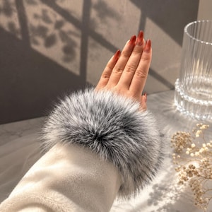 May include: A close-up of a white sleeve with a fluffy, gray and white fur cuff. The hand has long, red-orange nails. The background is blurred, with a glass and dried flowers visible.