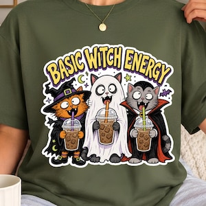 May include: An olive green t-shirt featuring a graphic of three cats in Halloween costumes holding iced coffee drinks. The text above reads BASIC WITCH ENERGY in yellow letters. The cats are dressed as a witch, a ghost, and a vampire.