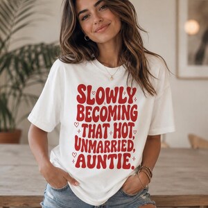 May include: A white short-sleeved t-shirt featuring red text that reads SLOWLY BECOMING THAT HOT, UNMARRIED, AUNTIE. The design includes small red heart and star icons surrounding the lettering, printed on a casual crew neck cotton shirt.