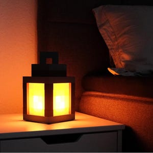 May include: A black, cube-shaped lantern with a handle, emitting a warm yellow glow. The lantern sits on a white nightstand next to a bed with a pillow. The scene is illuminated by the lantern's light, creating a cozy ambiance.