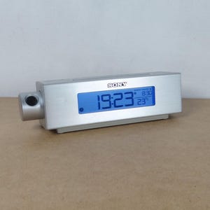 May include: A silver Sony digital alarm clock with a rectangular brushed metal finish. The front features a blue backlit LCD screen displaying the time 19:23, the temperature 23 degrees Celsius, and an alarm setting of 8:30. A circular projector lens is on the left side.