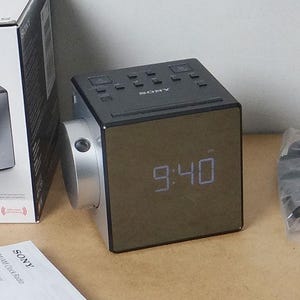 Puede incluir: Un despertador cúbico Sony ICF-C1PJ en negro y plata con una pantalla digital que muestra las 9:40, acompañado de su embalaje original de cartón, un montón de manuales de instrucciones y un pequeño accesorio negro sellado en una bolsa de plástico.