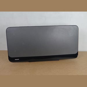 Peut inclure: Une enceinte Philips rectangulaire avec une façade en tissu gris foncé et une base noire brillante. L'appareil présente un design plat et épuré aux coins arrondis, avec la marque Philips imprimée en blanc dans le coin inférieur gauche du cadre.