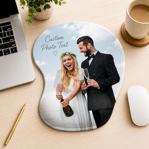 Op de afbeelding: Een gepersonaliseerde muismat met een afgedrukte foto van een persoon in een witte jurk die een champagnefles vasthoudt en een persoon in een zwart pak met een champagneglas, met de tekst Custom Photo Text en een hartje erboven.