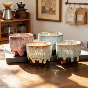 Könnte beinhalten: Vier Keramikbecher mit reaktiver Glasur. Ein Becher hat ein rosa-cremefarbenes Tropfmuster, einer ist hellblau-cremefarben und zwei haben cremefarbene und blaue Tropfen mit den in weißer Schreibschrift aufgedruckten Namen Nova und Mia.