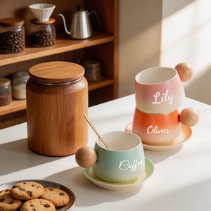 May include: Three ceramic mugs with round wooden handles and matching saucers. The mugs feature gradient colors and white text reading Lily, Oliver, and Coffee. A tall wooden canister with a lid and a plate of chocolate chip cookies are included.